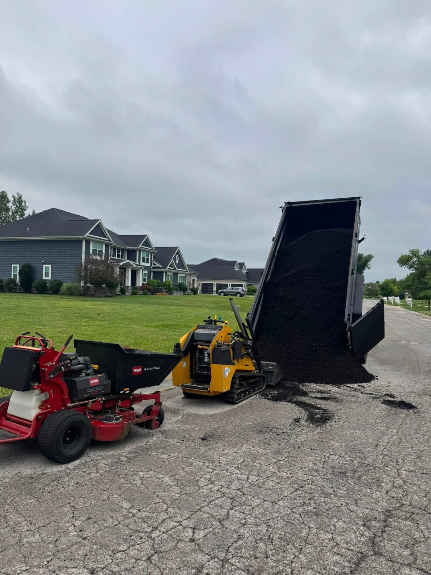 Dump trailer unloading black mulch beside a mulch buggy in a residential neighborhood